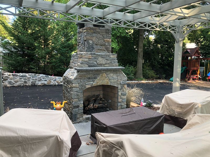 A stone fireplace is sitting under a pergola in a backyard.