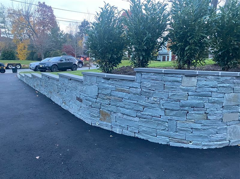 A stone wall surrounds a driveway with cars parked in front of it.