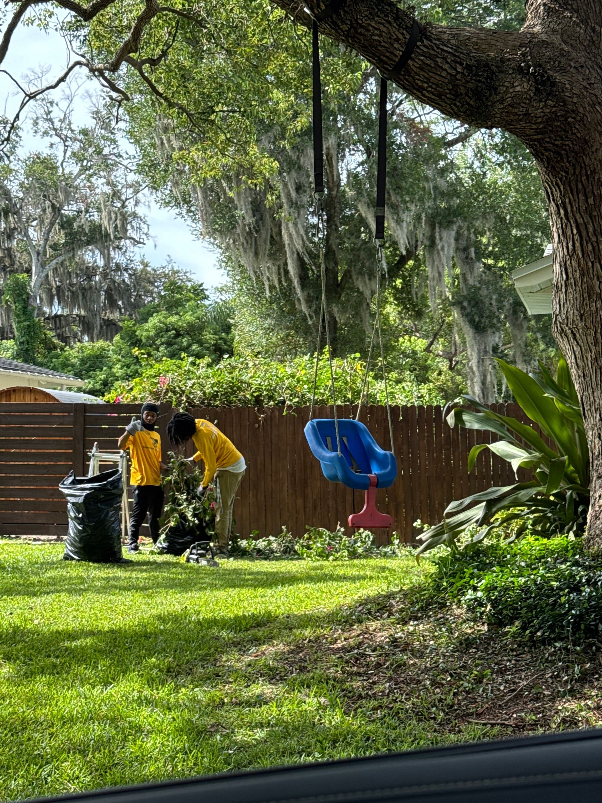 Two people in yellow shirts trimming yard, fence and swing set visible.