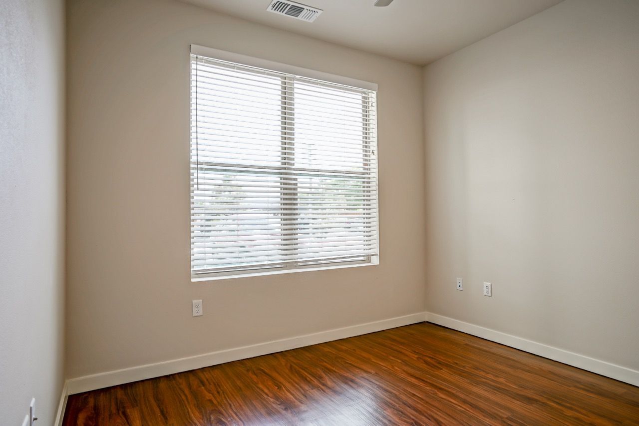 Empty bedroom with a large window, blinds, and wood floors.