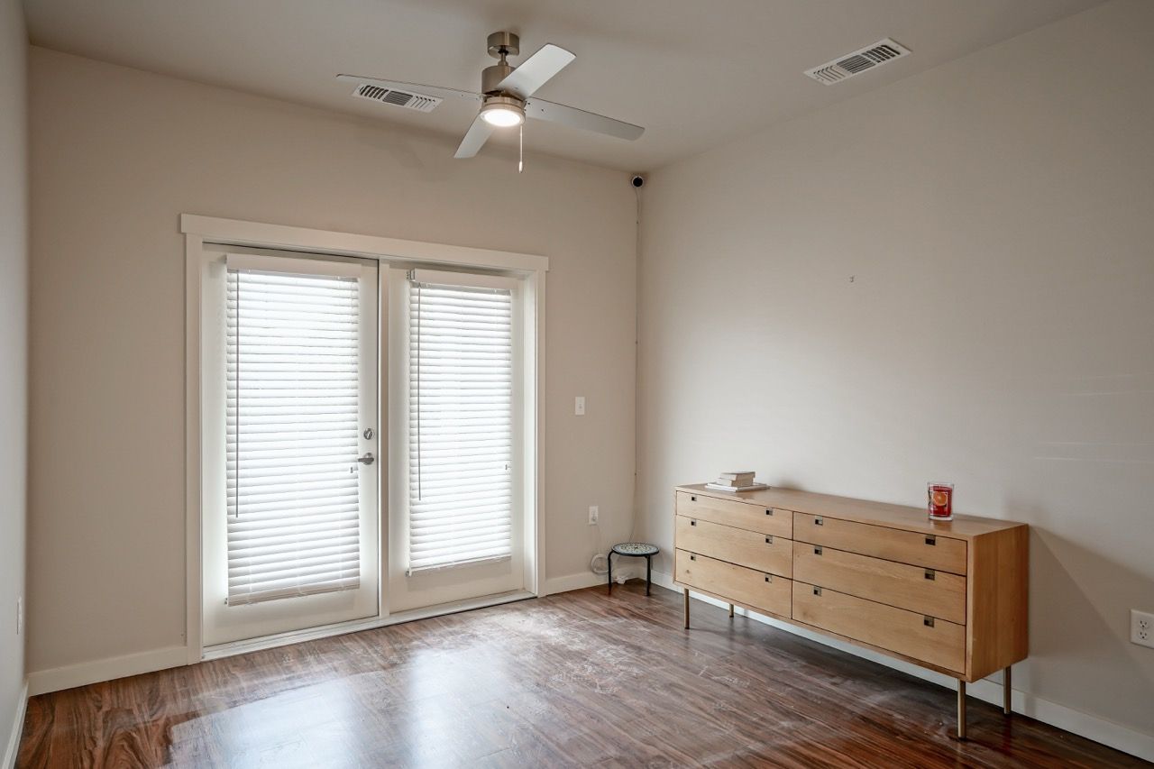Empty apartment bedroom with French doors, blinds, ceiling fan, and a wooden dresser.