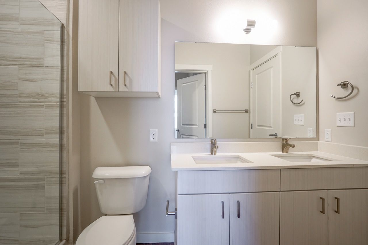 In-unit bathroom with double-sink vanity, large mirror, toilet, and tiled shower.