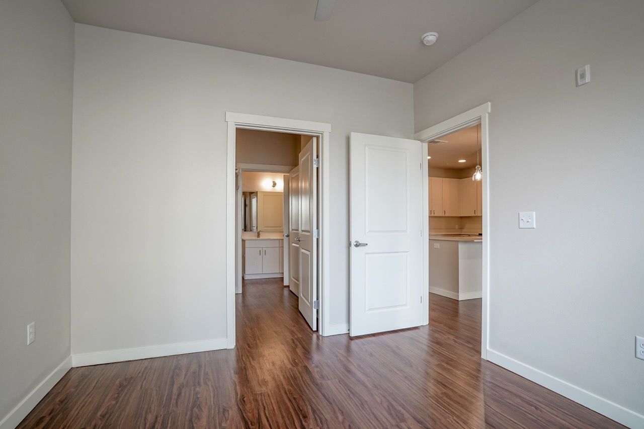 Empty apartment room with wood floor and open doors to the kitchen and bathroom.