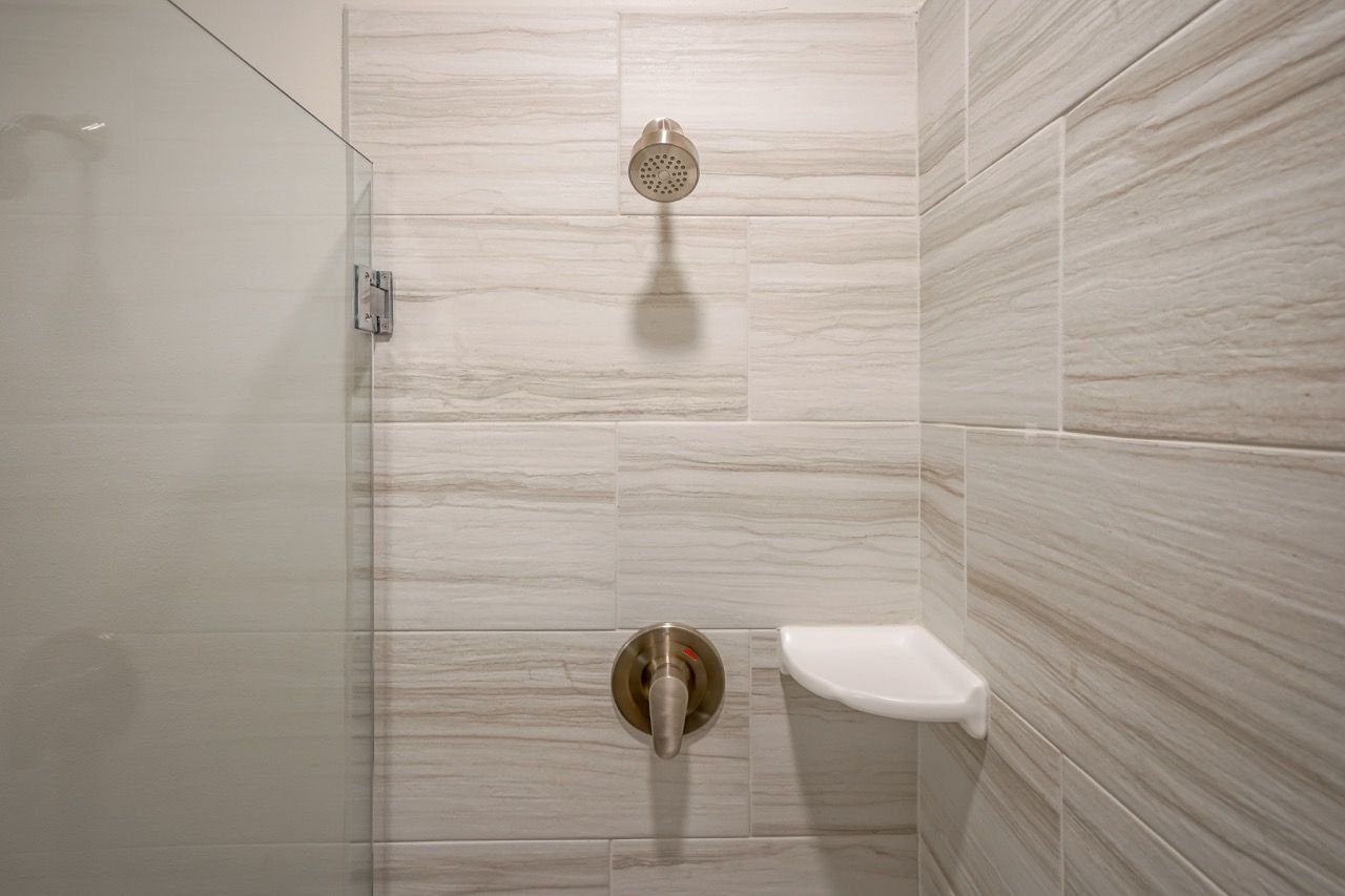 Shower area in a tiled bathroom with a glass door, ceiling-mounted showerhead, and a small corner shelf.