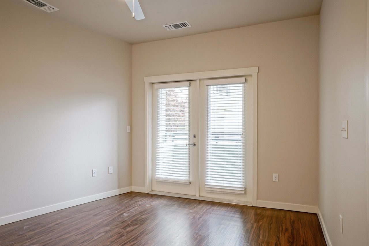 Empty living room with French doors and blinds, light beige walls, wooden floor.