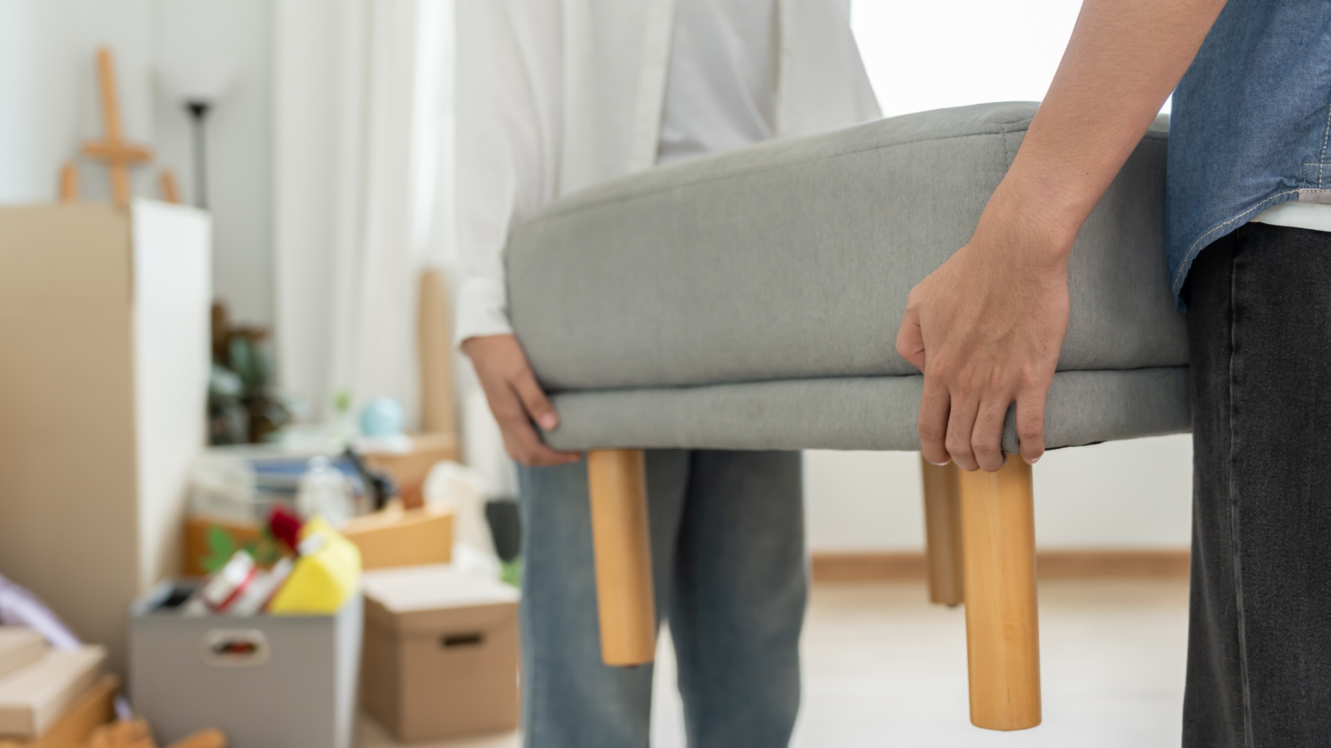 Two people lifting a gray upholstered ottoman with wooden legs in a room with moving boxes.