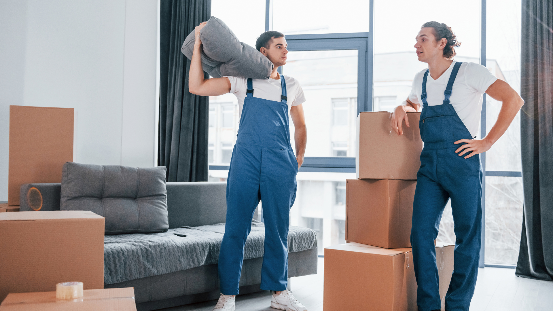 A man is carrying a pillow on his head while another man stands next to a pile of cardboard boxes.