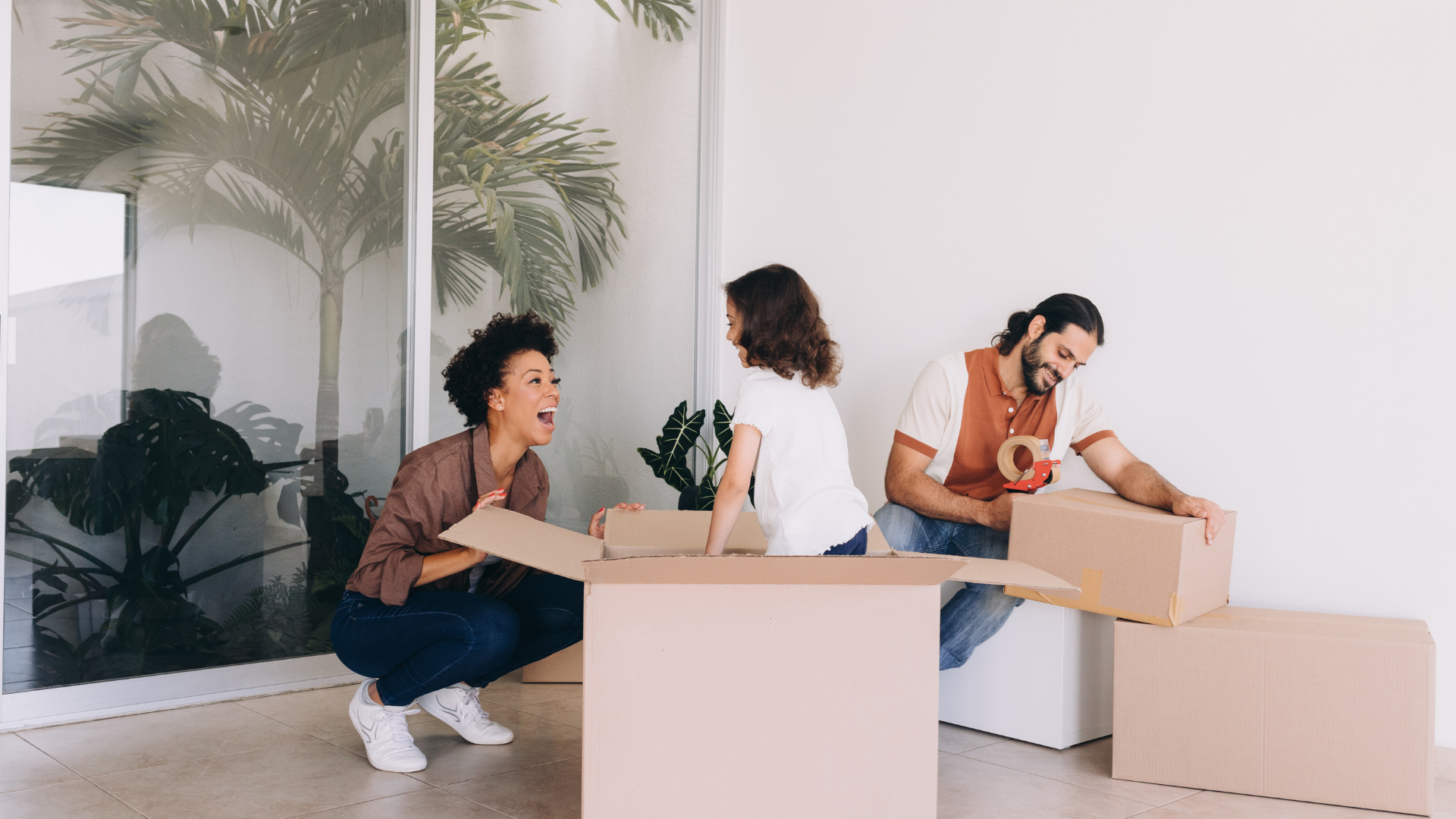 Three people unpacking boxes in a brightly lit room with plants. Two women and a man smiling.