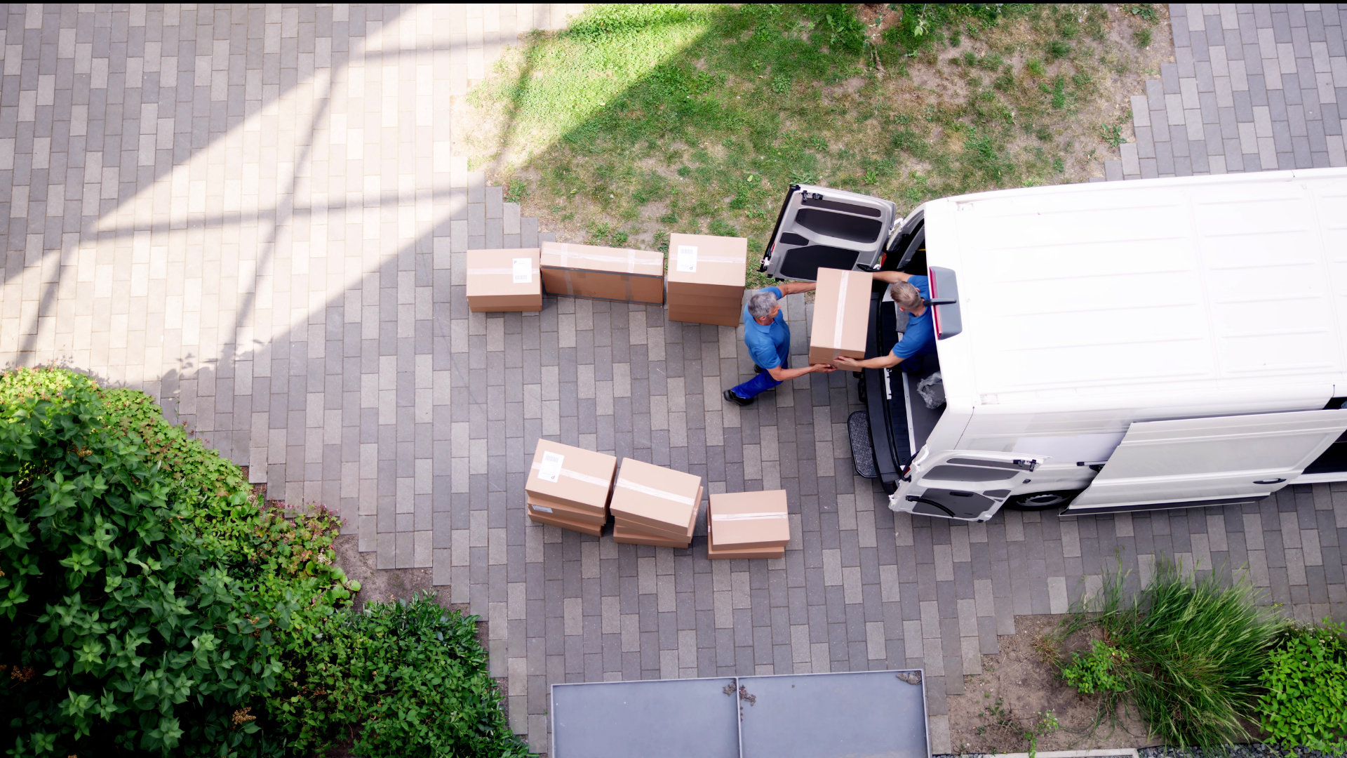 Two movers loading cardboard boxes into a white truck on a brick paved area.