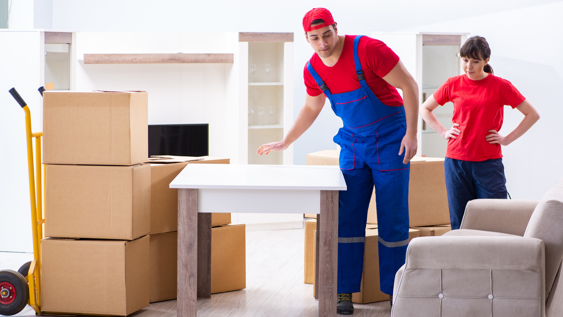 Movers in red and blue uniforms in a room, setting up a table. Boxes and furniture are present.