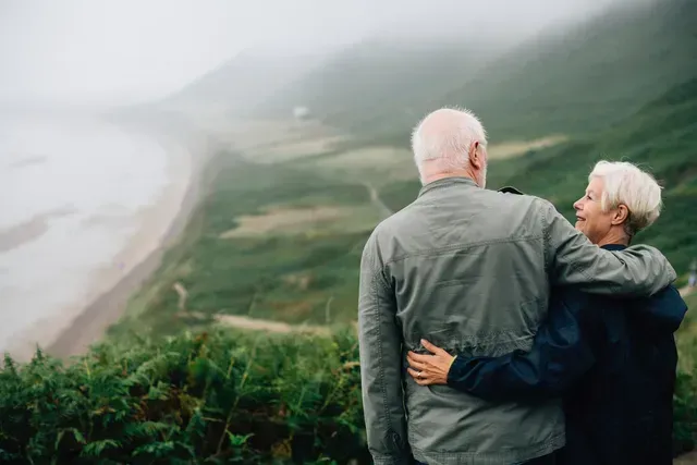 An elderly couple is standing on top of a hill looking at the ocean.