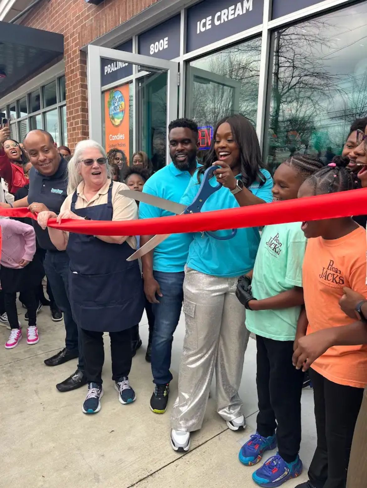 A group of people are cutting a red ribbon in front of an ice cream shop.