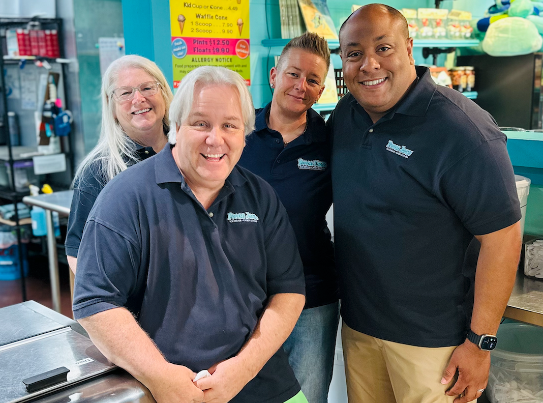Four people smiling together inside an ice cream shop. They wear matching blue shirts, counter visible.