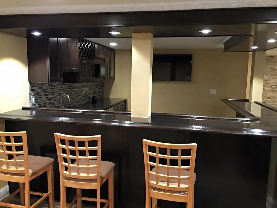 Dark wood bar with bar stools, cabinets, and a microwave in a basement setting.