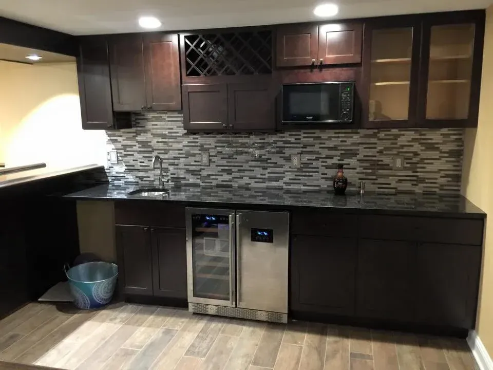 Dark wood bar with granite countertop, built-in wine cooler, microwave, wine rack, and sink. Mosaic backsplash.