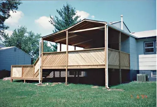 Wooden deck with covered roof attached to a light blue house, surrounded by green grass and trees.