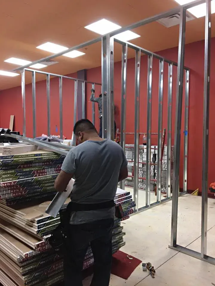 Man constructing metal framing for a wall indoors, with supplies nearby; red wall in background.