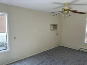 Empty room with gray carpet, beige walls, ceiling fan, window AC unit, and windows.