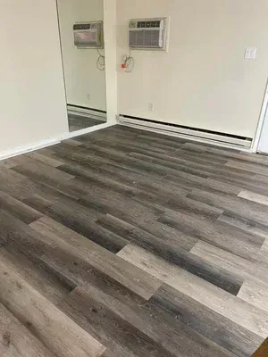 Empty room with grey wood-look flooring, white walls, mirror, and two air conditioners.