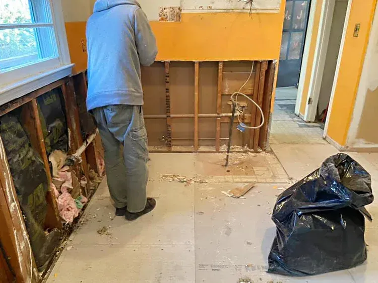 Person standing in a room under renovation, walls exposed, debris on floor, trash bag nearby.