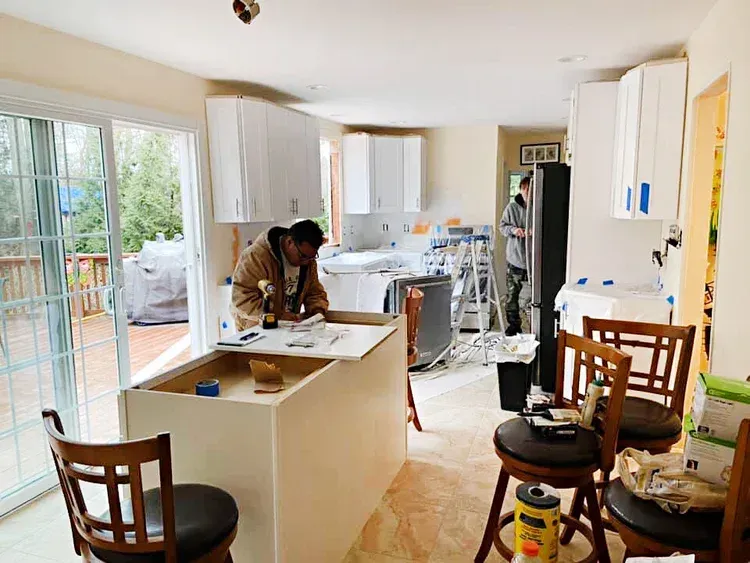 Kitchen renovation in progress: two workers install cabinets, island base. White and light wood interior, open to deck.