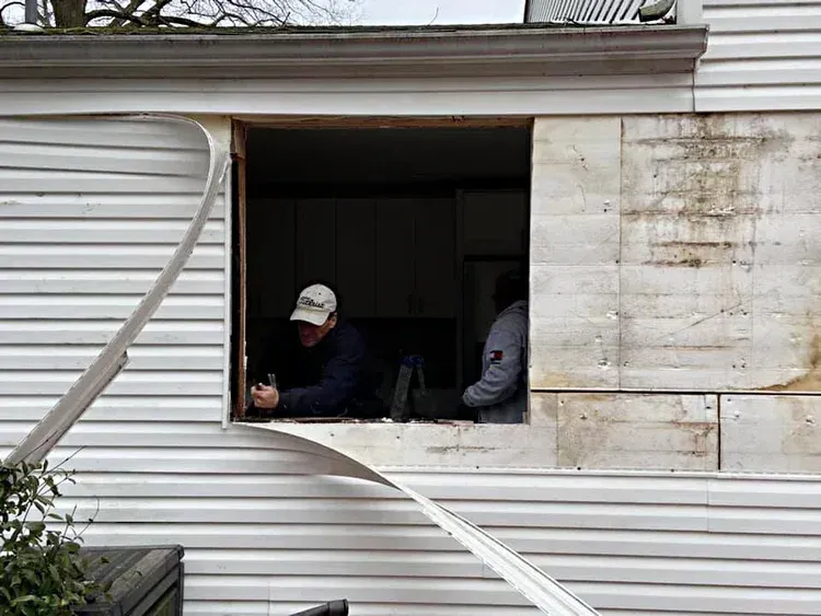 Exterior view of a mobile home window opening with two people inside, peeling away siding.