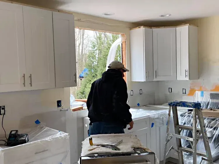 A person working in a kitchen with white cabinets, near a window. Construction in progress.