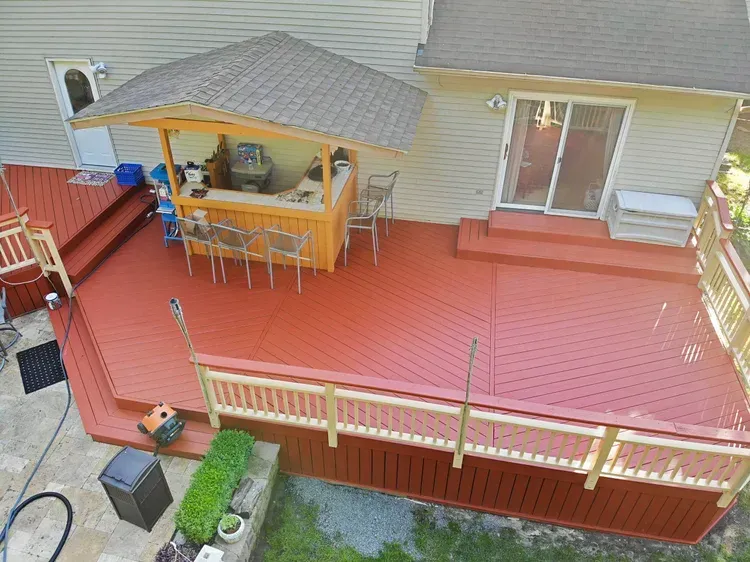 Red wooden deck with bar, railing, and steps, attached to a light-colored house.