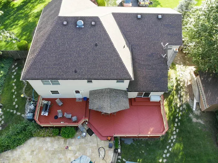 Aerial view of a two-story house with a brown roof and a red deck. Green lawn surrounds the house.