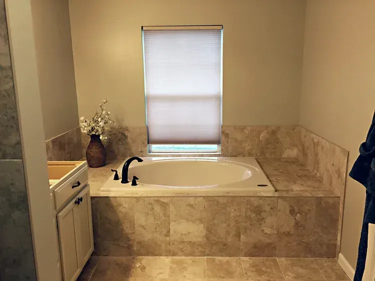 Bathroom with oval tub, beige tile surround, window with shade. Includes vanity and a dark faucet.