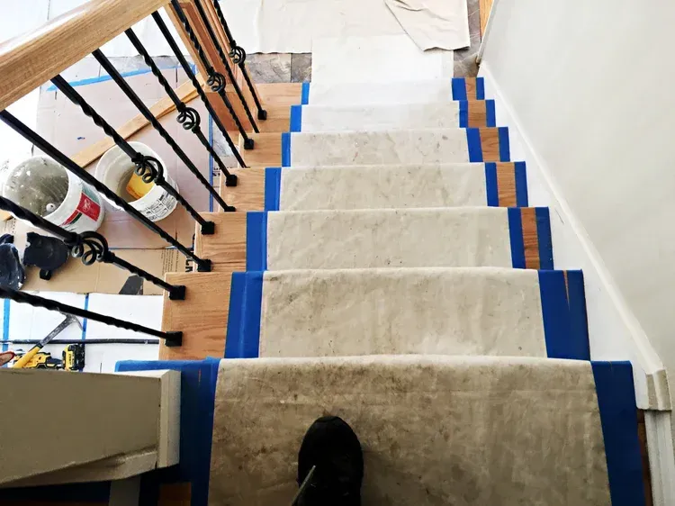 Staircase under renovation with black iron spindles, wooden handrail, and paper covering steps with blue tape.