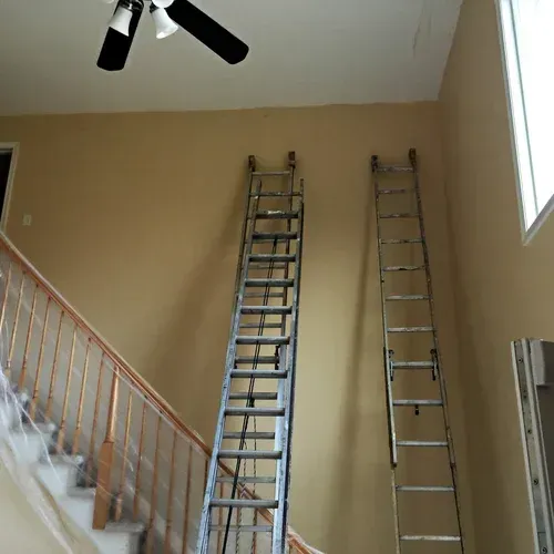 Two ladders leaning against a tall tan wall; staircase on the left, ceiling fan overhead.