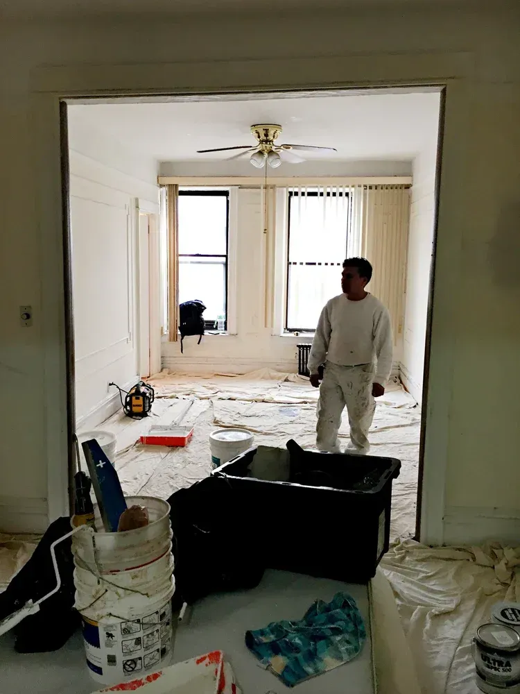 Man in paint-splattered overalls stands in a room under renovation, paint buckets and tools scattered around.