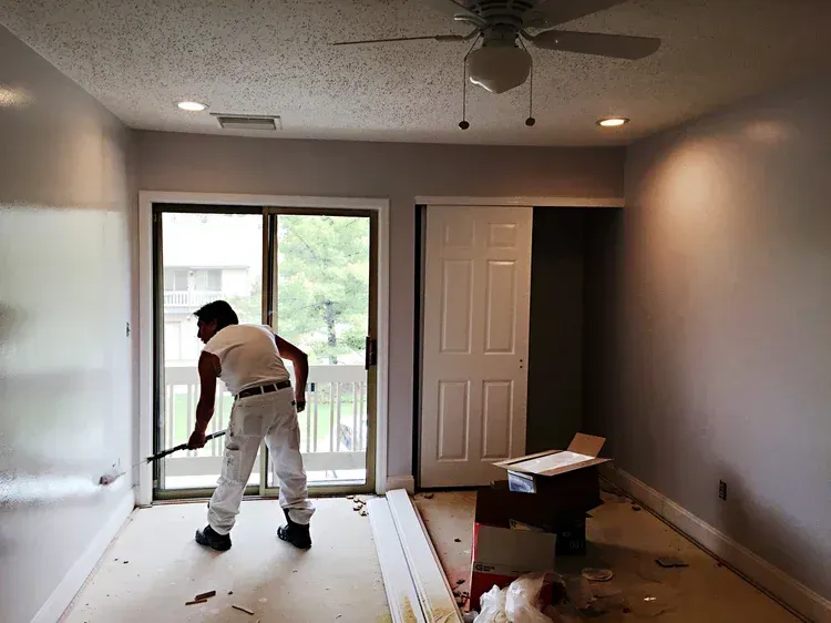 Painter in white overalls working on a room with gray walls, sliding glass door, and white trim.