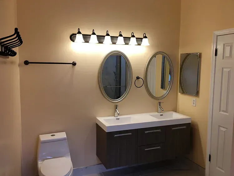 Bathroom with double sinks, two mirrors, and a toilet. Tan walls, gray vanity, and dark fixtures.