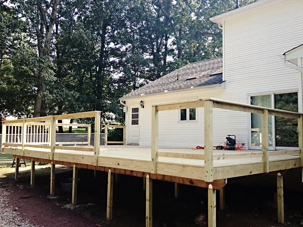 Wooden deck addition to a white house with a partially completed railing, surrounded by trees.