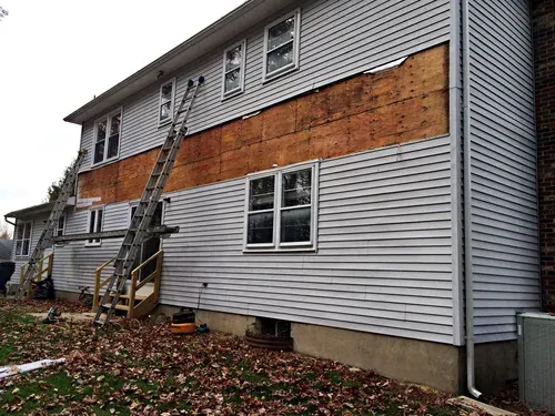 House siding removed, ladders in place. Exposed wood panels, gray siding below and above. Cloudy day.