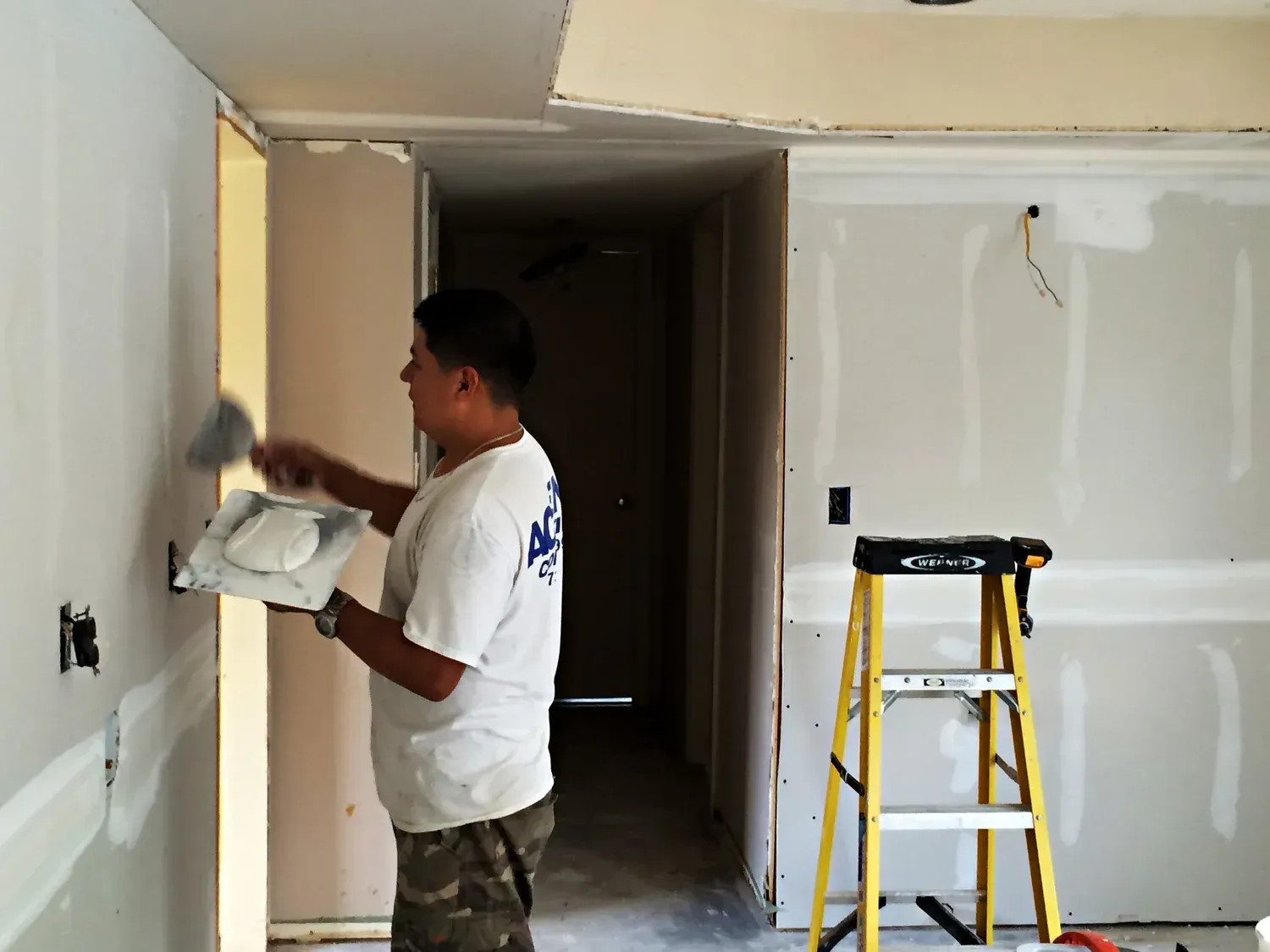 Man applying drywall mud with a trowel on a wall in a room under construction. A ladder is nearby.