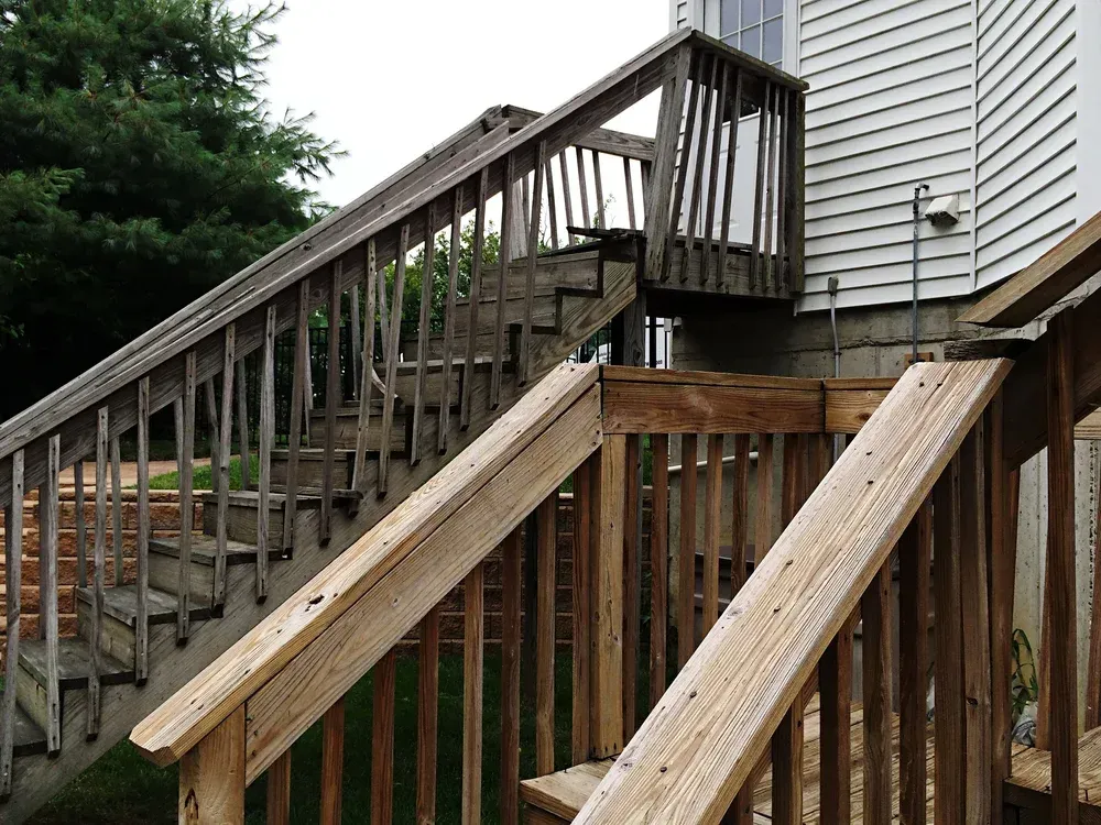 Wooden outdoor staircase and deck attached to a house with white siding; weathered.