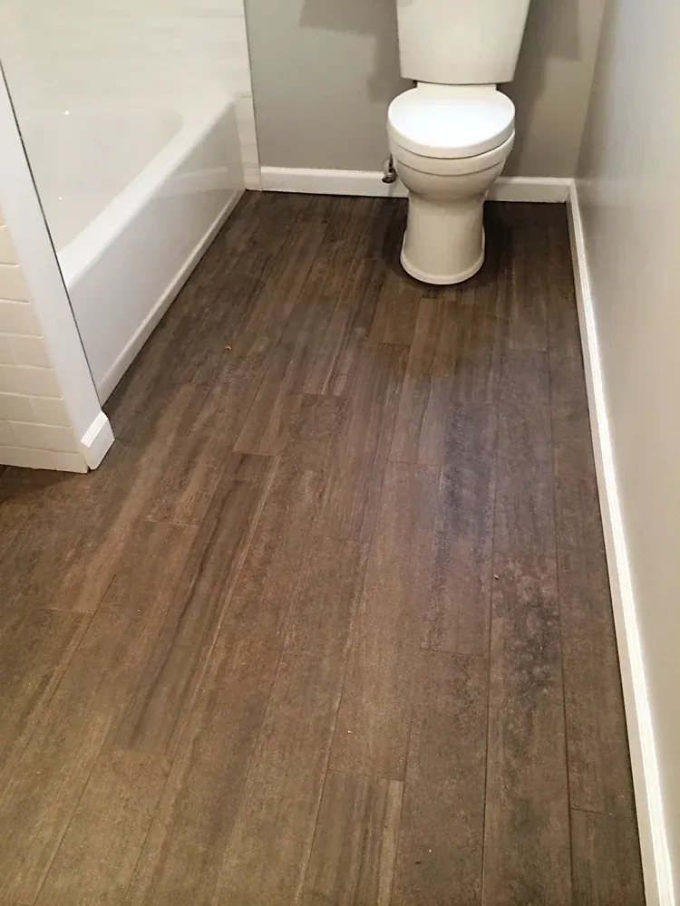 Bathroom with wood-look tile flooring. White tub and toilet. Gray walls, white baseboards.