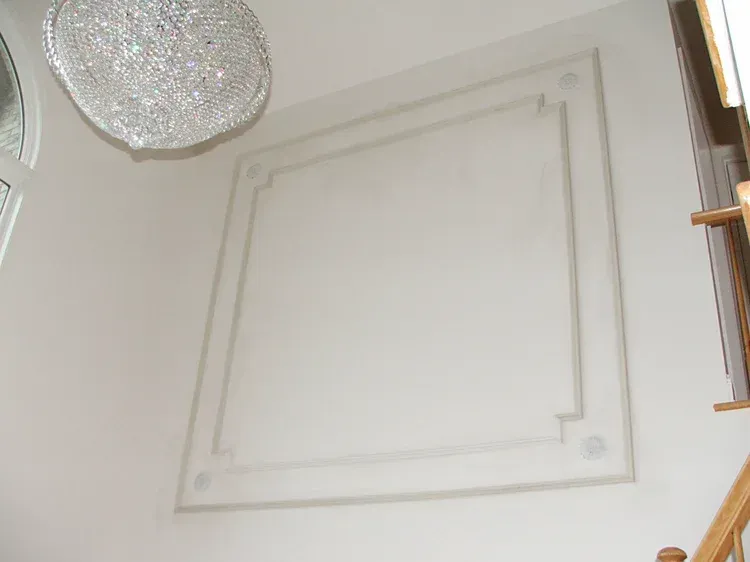 White wall with square decorative molding, crystal chandelier, and wooden staircase.
