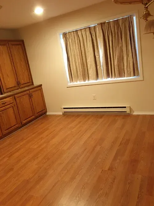 Empty room with wood-look flooring, cabinets, window with curtains, and a baseboard heater.