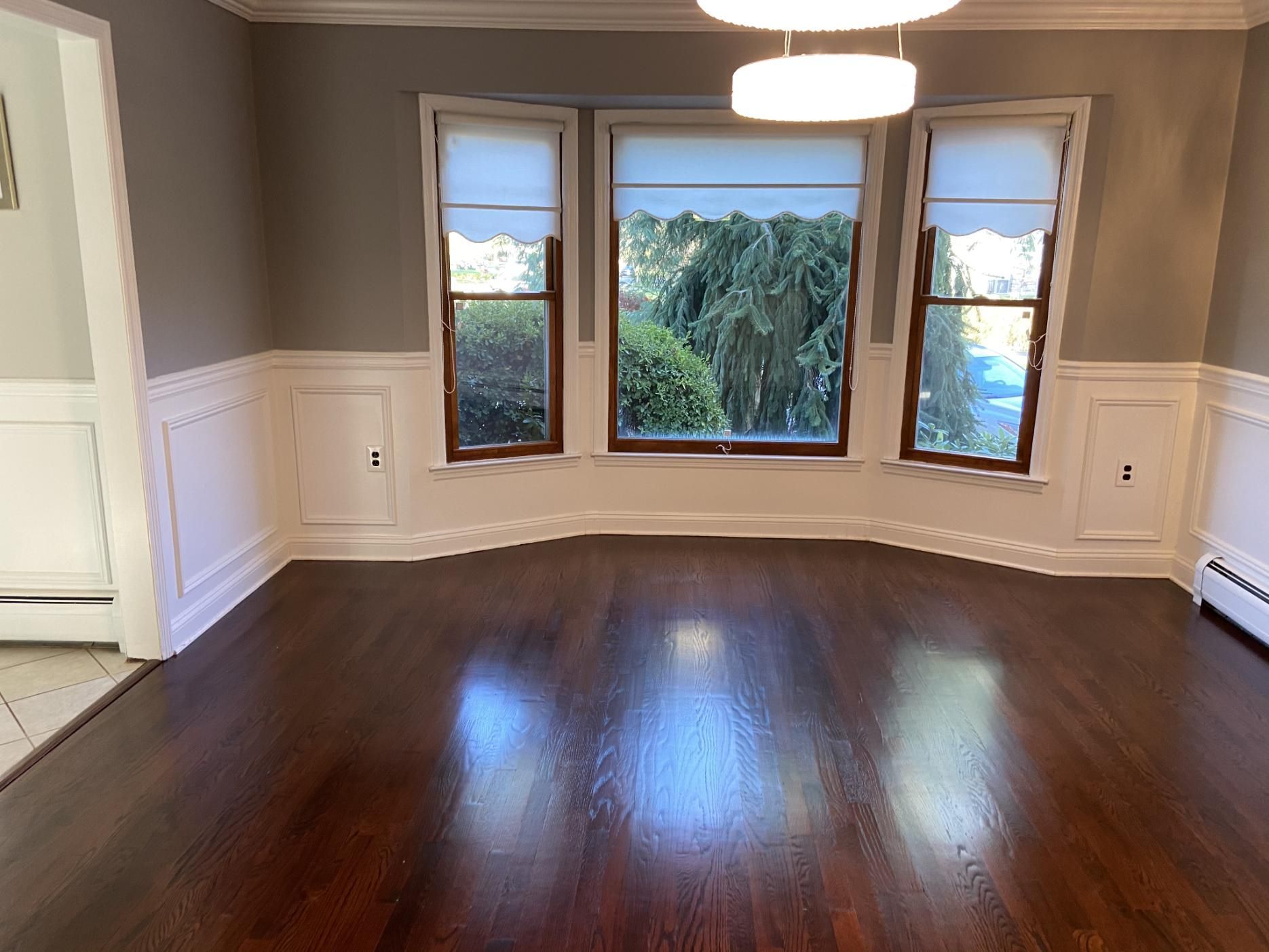 Empty dining room with dark wood floor, windows with shades, and white wainscoting.
