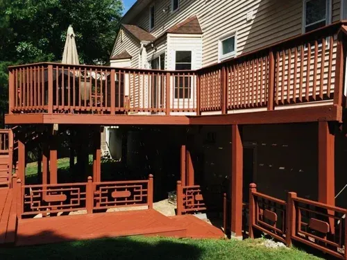 Two-level wooden deck attached to a house with brown paint. A set of stairs lead down from the lower level to the yard.
