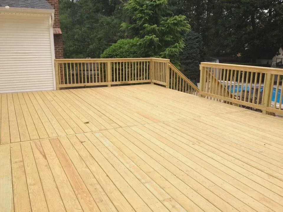 Wooden deck with railing, connected to a house. Trees and a pool visible in the background.