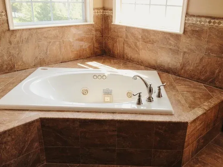 Corner bathtub surrounded by brown tile, next to a window.