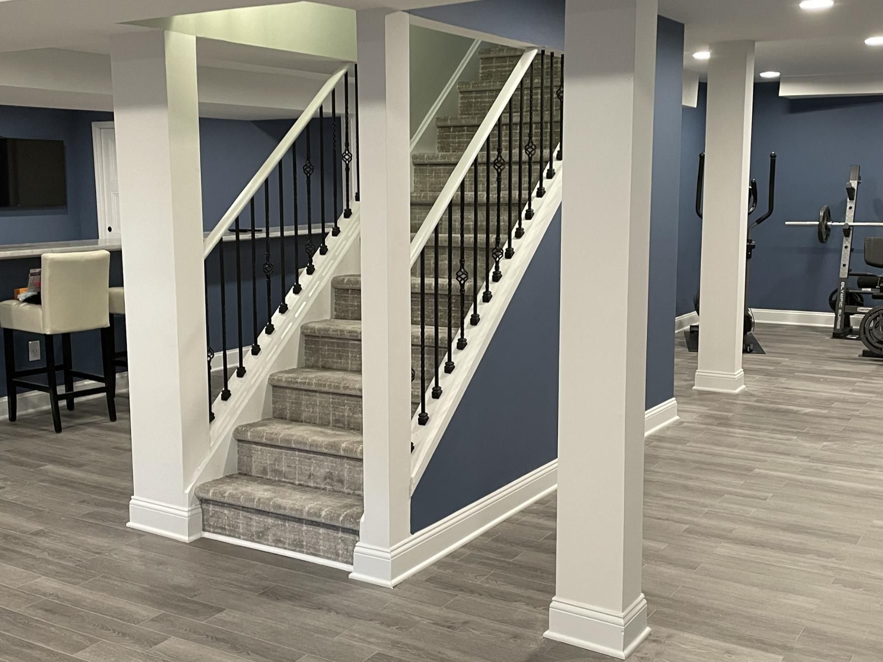 Basement with staircase, gray floors, white columns, blue walls, black railing, and fitness equipment.