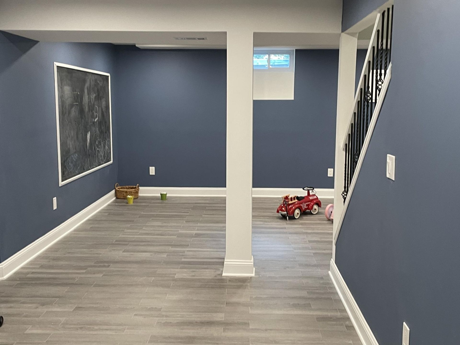 Finished basement with gray flooring, blue walls, white trim, and a chalkboard. A toy car and basket sit on the floor.