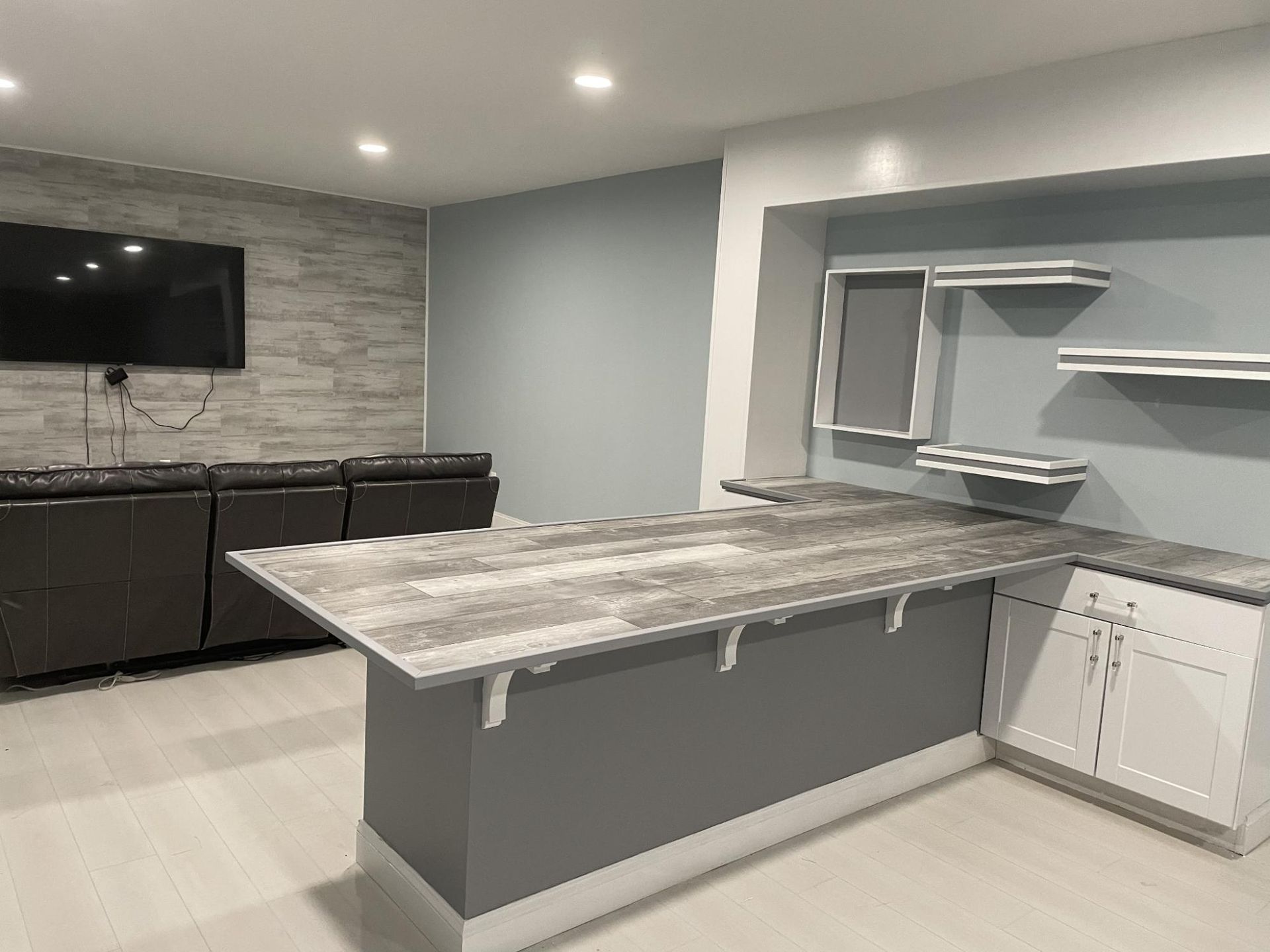 Basement bar area with gray and white color scheme. Dark gray couch and TV on wall.