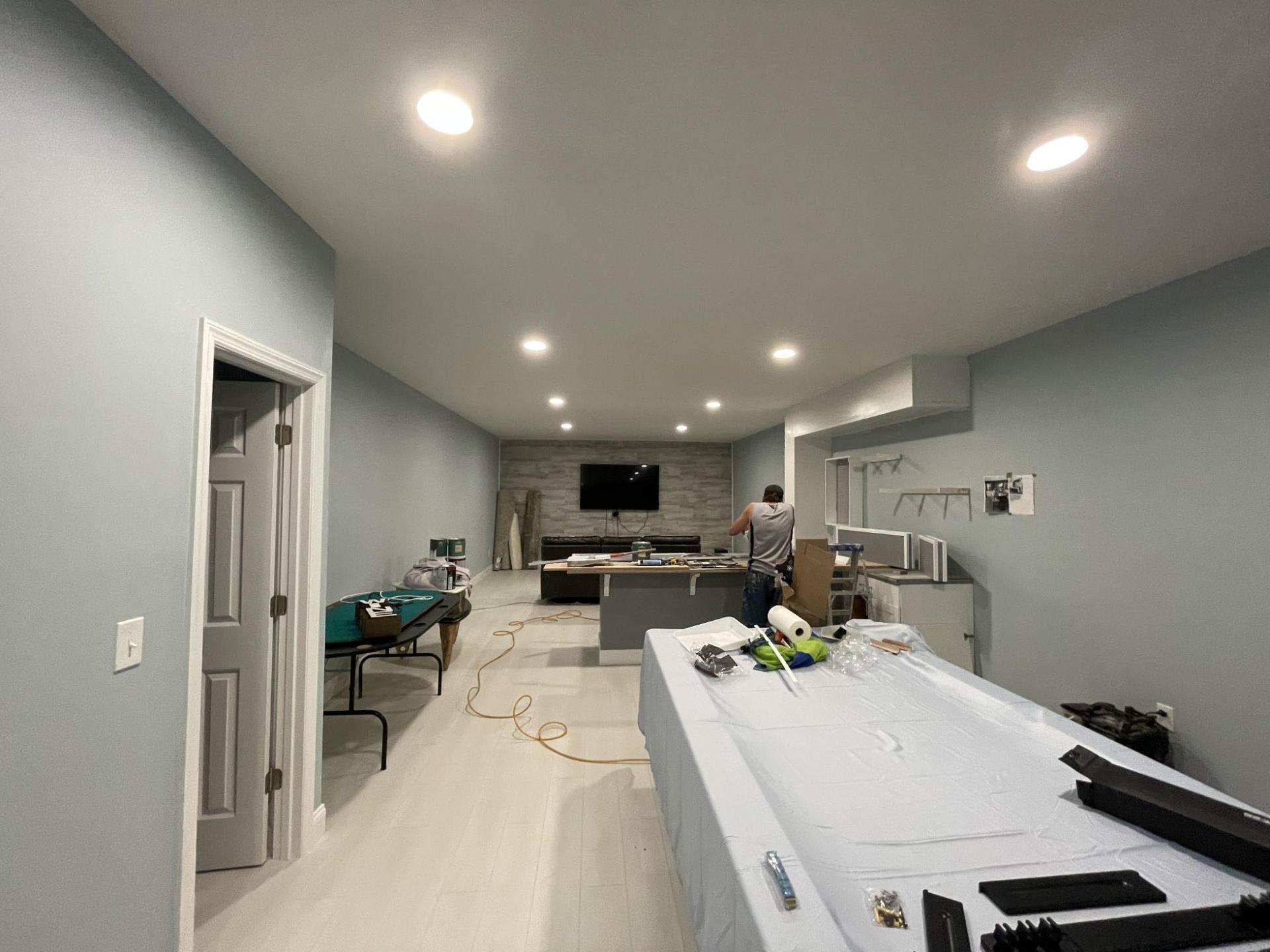 Spacious room under renovation: light blue walls, gray flooring, recessed lighting. Person working near gray counter and TV.